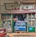 A small, colorful storefront selling groceries and household items. The entrance is filled with various products, including groceries in a blue display fridge. Shelves on either side showcase items like cooking oils and detergents. The store has a sign in a language that uses an Arabic script above the door.