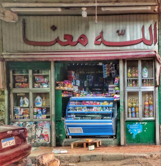 A small, colorful storefront selling groceries and household items. The entrance is filled with various products, including groceries in a blue display fridge. Shelves on either side showcase items like cooking oils and detergents. The store has a sign in a language that uses an Arabic script above the door.
