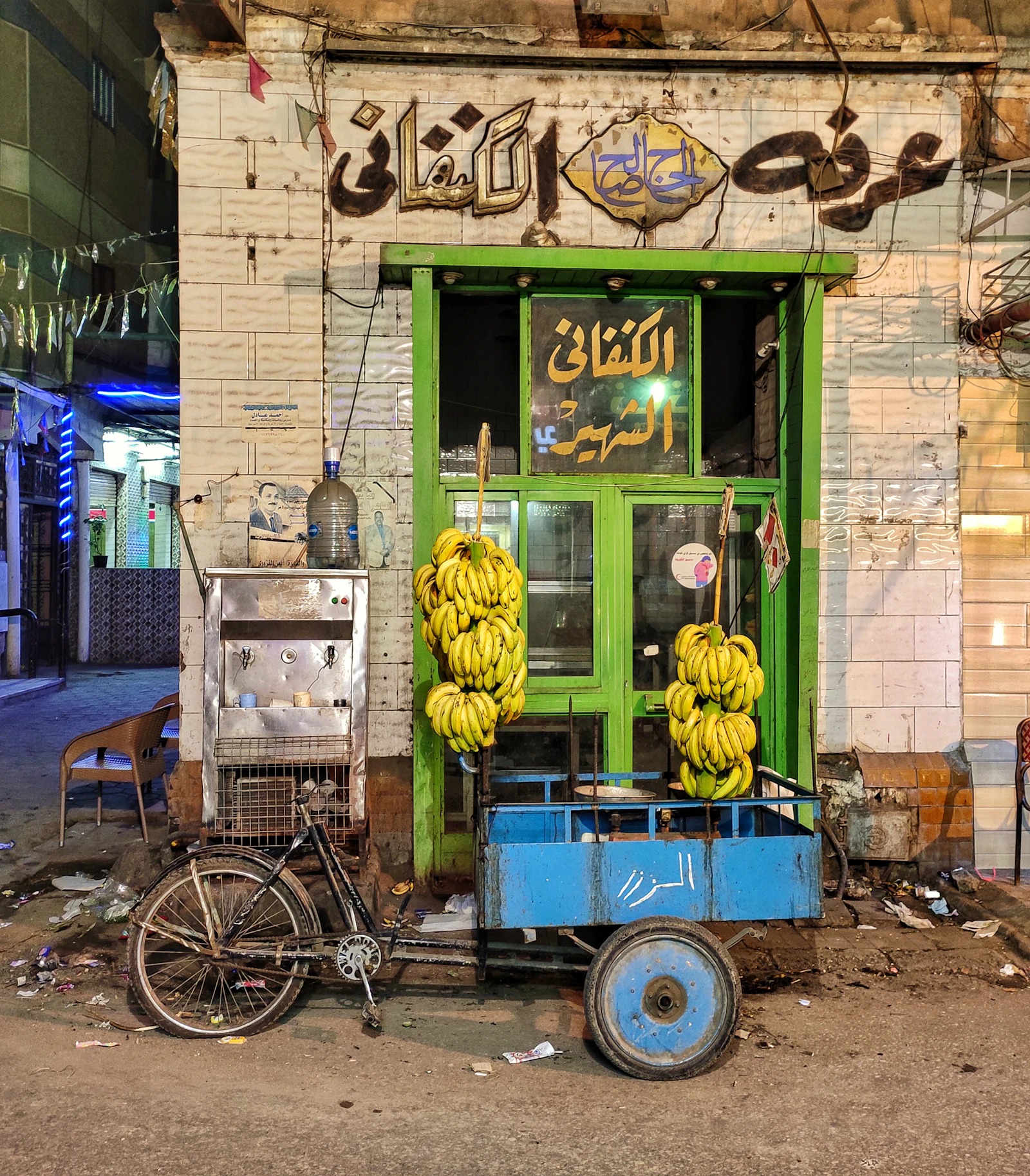 Illustrative image for vegan food in cairo: ful, taameya and koshari: Large bunches of bananas hanging from a blue bicycle cart outside a green Cairo shopfront.