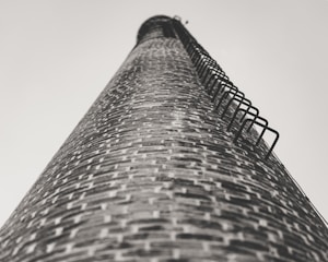 A tall brick chimney stretches skyward, captured from a low angle. The brickwork is detailed, with metal rungs attached along the side for climbing. The perspective creates a sense of height and scale.
