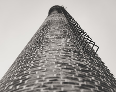 A tall brick chimney stretches skyward, captured from a low angle. The brickwork is detailed, with metal rungs attached along the side for climbing. The perspective creates a sense of height and scale.