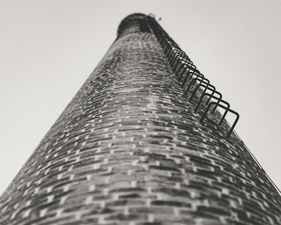 A tall brick chimney stretches skyward, captured from a low angle. The brickwork is detailed, with metal rungs attached along the side for climbing. The perspective creates a sense of height and scale.