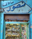 An old shop facade with Arabic writing at the top. The storefront display is filled with an assortment of wristwatches and various small, colorful boxes or packaging. The exterior has peeling blue paint, giving it a vintage and worn-out appearance.