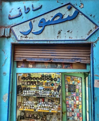 An old shop facade with Arabic writing at the top. The storefront display is filled with an assortment of wristwatches and various small, colorful boxes or packaging. The exterior has peeling blue paint, giving it a vintage and worn-out appearance.