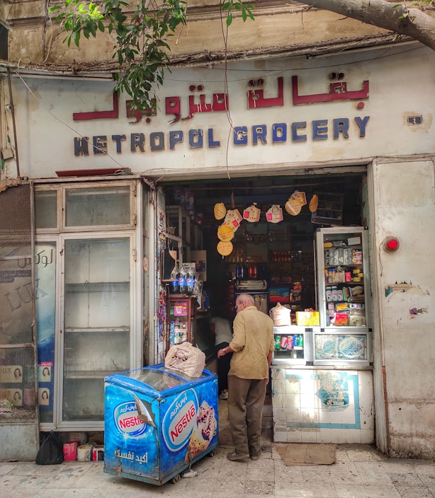 A small, local grocery store with a visible sign reading 'Metropol Grocery' in both English and Arabic. The storefront displays a variety of products, including items on shelves and a Nestle-branded refrigerator. A man is standing near the entrance with a bag, facing towards the inside of the store. The exterior shows signs of age and wear, with a tree branch partially in view above.