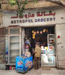 A small, local grocery store with a visible sign reading 'Metropol Grocery' in both English and Arabic. The storefront displays a variety of products, including items on shelves and a Nestle-branded refrigerator. A man is standing near the entrance with a bag, facing towards the inside of the store. The exterior shows signs of age and wear, with a tree branch partially in view above.