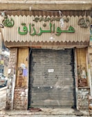 A closed storefront with metal shutters, surrounded by stone-patterned walls. The sign above the entrance features Arabic script in large green letters. The shop appears to be located on a narrow street with a parked car partially visible on the left. The exterior shows signs of wear and age, with peeled paint and weathered surfaces.