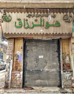 A closed storefront with metal shutters, surrounded by stone-patterned walls. The sign above the entrance features Arabic script in large green letters. The shop appears to be located on a narrow street with a parked car partially visible on the left. The exterior shows signs of wear and age, with peeled paint and weathered surfaces.