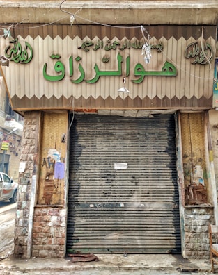 A closed storefront with metal shutters, surrounded by stone-patterned walls. The sign above the entrance features Arabic script in large green letters. The shop appears to be located on a narrow street with a parked car partially visible on the left. The exterior shows signs of wear and age, with peeled paint and weathered surfaces.