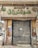 A closed storefront with metal shutters, surrounded by stone-patterned walls. The sign above the entrance features Arabic script in large green letters. The shop appears to be located on a narrow street with a parked car partially visible on the left. The exterior shows signs of wear and age, with peeled paint and weathered surfaces.