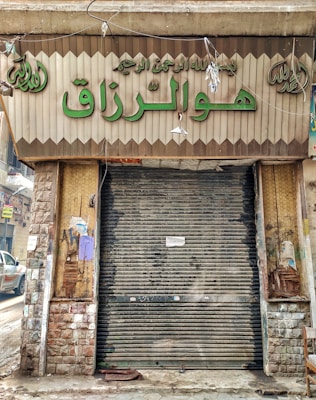 A closed storefront with metal shutters, surrounded by stone-patterned walls. The sign above the entrance features Arabic script in large green letters. The shop appears to be located on a narrow street with a parked car partially visible on the left. The exterior shows signs of wear and age, with peeled paint and weathered surfaces.