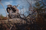 An off-road motorcycle is parked on a rocky terrain surrounded by dry branches and sparse vegetation. The bike features knobby tires and gear designed for rugged use. The sky is partly cloudy, suggesting an adventurous outdoor setting.