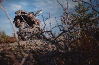 An off-road motorcycle is parked on a rocky terrain surrounded by dry branches and sparse vegetation. The bike features knobby tires and gear designed for rugged use. The sky is partly cloudy, suggesting an adventurous outdoor setting.