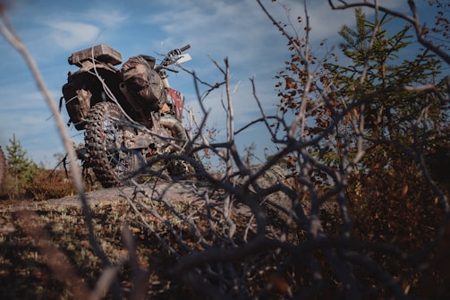An off-road motorcycle is parked on a rocky terrain surrounded by dry branches and sparse vegetation. The bike features knobby tires and gear designed for rugged use. The sky is partly cloudy, suggesting an adventurous outdoor setting.