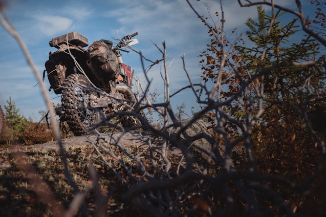 An off-road motorcycle is parked on a rocky terrain surrounded by dry branches and sparse vegetation. The bike features knobby tires and gear designed for rugged use. The sky is partly cloudy, suggesting an adventurous outdoor setting.