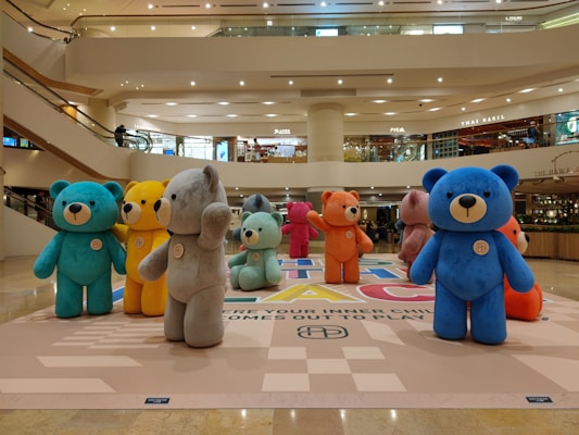 A group of large, colorful teddy bear statues are displayed in a shopping mall. They are arranged in a semicircle on a patterned floor, with surrounding balconies and stores visible in the background. Each bear is made of a soft material and has a unique color, including blue, orange, yellow, gray, and pink.
