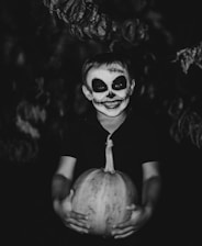 A child with face paint resembling a skull smiles while holding a pumpkin. The background is dark, enhancing the spooky and Halloween-themed atmosphere.