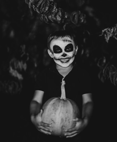 A child with face paint resembling a skull smiles while holding a pumpkin. The background is dark, enhancing the spooky and Halloween-themed atmosphere.