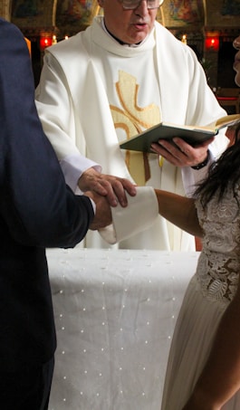 A person in a religious robe is officiating a ceremony, holding a book while another individual places their hand over a third person's hand. The setting appears to be a church.