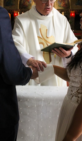 A person in a religious robe is officiating a ceremony, holding a book while another individual places their hand over a third person's hand. The setting appears to be a church.
