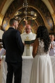 A couple stands before a priest in a church, likely during a wedding ceremony. They are facing the priest, who is holding a book and wearing a white robe with a gold cross. The church features an ornate and colorful mosaic backdrop with arched designs and a chandelier above.
