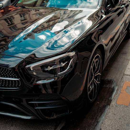A close-up view of a black luxury car parked on a city street. The photo highlights the car's sleek design and glossy finish, reflecting buildings and sky on its surface. The image showcases the front part of the car, including the headlights, the grille, and part of the wheels.