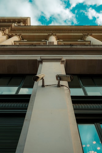 A low-angle view of a building facade featuring two surveillance cameras mounted on a central pillar. The architectural design includes ornate details with columns and decorative moldings. The sky is partly cloudy in the background, providing a contrast to the building's beige and gray tones.