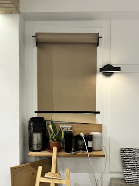 A small wooden shelf is mounted on a white wall beneath a rolled-up brown shade. On the shelf, there are various items including a potted plant, a black cylindrical lamp, a coffee tamper, bottles, and a cylindrical light or speaker. An easel leans against the wall nearby. The setting appears to be cozy and organized.