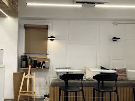 A cozy indoor space with a minimalistic design. The room features a bench with multiple cushions, two small round tables, and black chairs. A wooden structure resembling a small ladder or stand is positioned next to a shelf, which holds books, a potted plant, and some small decorative items. There's a window with a rolled-down beige blind, and the wall is tastefully decorated with panel designs and modern light fixtures. The lighting is warm, creating an inviting atmosphere.