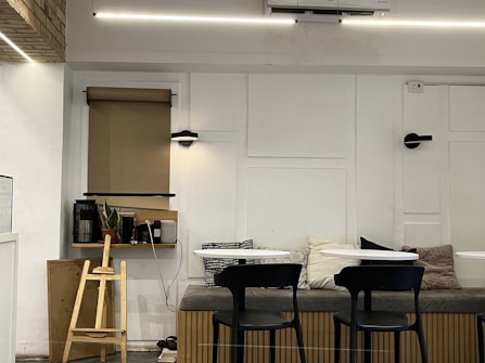 A cozy indoor space with a minimalistic design. The room features a bench with multiple cushions, two small round tables, and black chairs. A wooden structure resembling a small ladder or stand is positioned next to a shelf, which holds books, a potted plant, and some small decorative items. There's a window with a rolled-down beige blind, and the wall is tastefully decorated with panel designs and modern light fixtures. The lighting is warm, creating an inviting atmosphere.