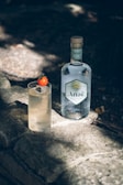 A bottle of Anaë gin stands on a stone surface next to a cocktail with ice cubes and a cherry tomato garnish. The scene is outdoors, under soft natural lighting.