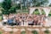A large group of people is gathered together outdoors, likely for a celebratory occasion such as a wedding. The group includes individuals of various ages, some in formal attire including dresses and suits. The background features lush greenery, trees, a pergola with white drapes, and a well-maintained garden area. Many of the people are smiling and raising their hands in a celebratory gesture.