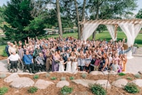 A large group of people is gathered together outdoors, likely for a celebratory occasion such as a wedding. The group includes individuals of various ages, some in formal attire including dresses and suits. The background features lush greenery, trees, a pergola with white drapes, and a well-maintained garden area. Many of the people are smiling and raising their hands in a celebratory gesture.