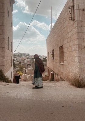 A person wearing a patterned long skirt and a dark headscarf walks down a narrow street between two stone buildings. Another person is seen in the distance, also walking away. The buildings have a rustic feel with visible wires and a slightly cloudy sky above. In the background, a town with closely packed buildings is visible.