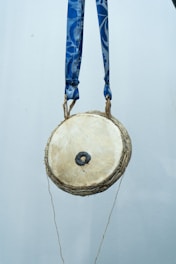 A small, round traditional drum hangs suspended by two blue straps. The drumhead is a light beige color with a dark central patch, likely used for tuning or sound variation. The background is plain and muted, highlighting the drum as the focal point of the image.