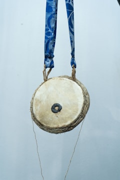 A small, round traditional drum hangs suspended by two blue straps. The drumhead is a light beige color with a dark central patch, likely used for tuning or sound variation. The background is plain and muted, highlighting the drum as the focal point of the image.