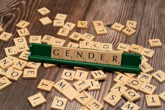 A green Scrabble tile holder displays the word 'GENDER' using wooden letter tiles. The surrounding area is scattered with additional Scrabble tiles on a wooden surface.