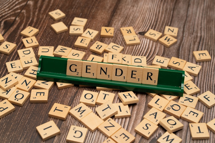 A green Scrabble tile holder displays the word 'GENDER' using wooden letter tiles. The surrounding area is scattered with additional Scrabble tiles on a wooden surface.