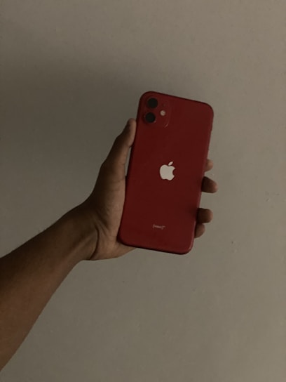 A hand is holding a red smartphone with a prominent apple logo on the back against a plain background. The phone has two camera lenses and a minimalist design.