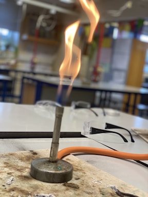 A Bunsen burner is on a lab table with a visible flame, surrounded by laboratory equipment like safety goggles. The lab room is well-lit, with some colorful elements in the background, possibly charts or materials on the wall.