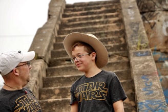 Two people, both wearing black Star Wars T-shirts, are standing in front of old stone steps covered with some graffiti. The younger one is wearing glasses and a cowboy hat, smiling slightly.