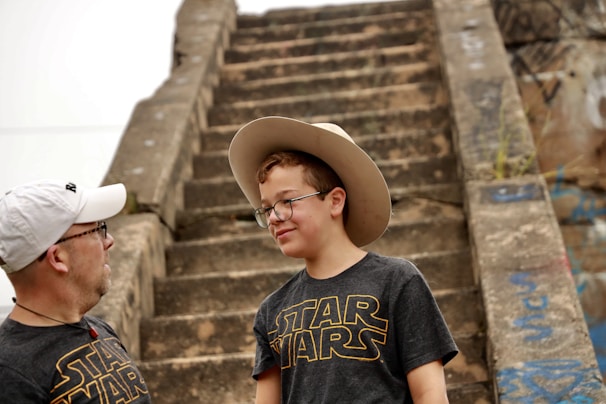 Two people, both wearing black Star Wars T-shirts, are standing in front of old stone steps covered with some graffiti. The younger one is wearing glasses and a cowboy hat, smiling slightly.