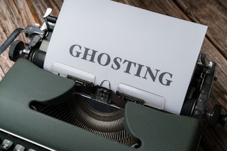 A vintage typewriter with a sheet of paper inserted showing the word 'GHOSTING' in bold letters. The typewriter is placed on a wooden surface, and the image has a retro aesthetic.