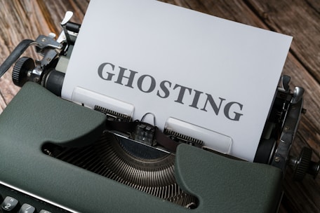 A vintage typewriter with a sheet of paper inserted showing the word 'GHOSTING' in bold letters. The typewriter is placed on a wooden surface, and the image has a retro aesthetic.