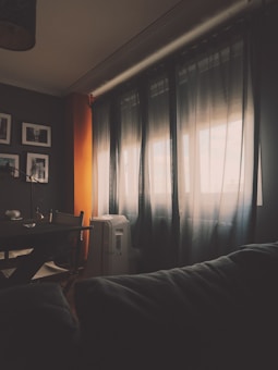 A dimly lit room features sheer curtains filtering light from a window, casting soft shadows across the space. A dark couch is visible in the foreground, while a table with chairs and an air conditioning unit stand near the window. The walls are adorned with framed photographs, and an orange accent wall adds warmth to the ambiance.