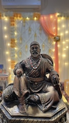 A bronze statue of a seated historical figure, adorned with a beaded necklace, in front of a decorative background featuring orange and white drapery with string lights. The statue is intricately detailed, depicting traditional attire.