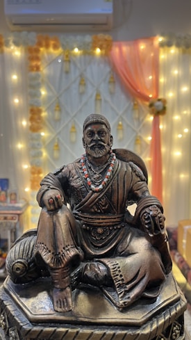 A bronze statue of a seated historical figure, adorned with a beaded necklace, in front of a decorative background featuring orange and white drapery with string lights. The statue is intricately detailed, depicting traditional attire.