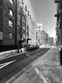 A black and white urban street scene featuring tall buildings with windows, a narrow street lined with cars, and sidewalks. The architecture combines a mix of traditional and modern elements. Some people are visible walking along the sidewalks, and bollards line the street, enhancing the perspective.