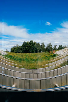 A modern landscape design featuring tiered grassy slopes outlined by sleek metal railings, with a backdrop of lush green trees and a clear blue sky above.