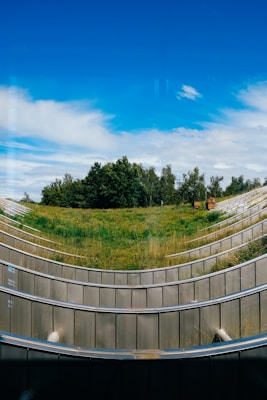 A modern landscape design featuring tiered grassy slopes outlined by sleek metal railings, with a backdrop of lush green trees and a clear blue sky above.